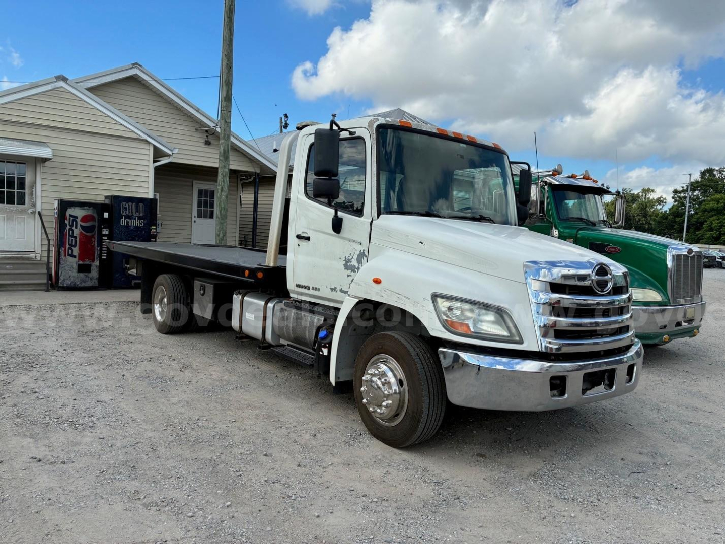 Hino conventional cab tow truck on gravel lot, white cab, chrome front bumper, flatbed service body, other green truck in background.