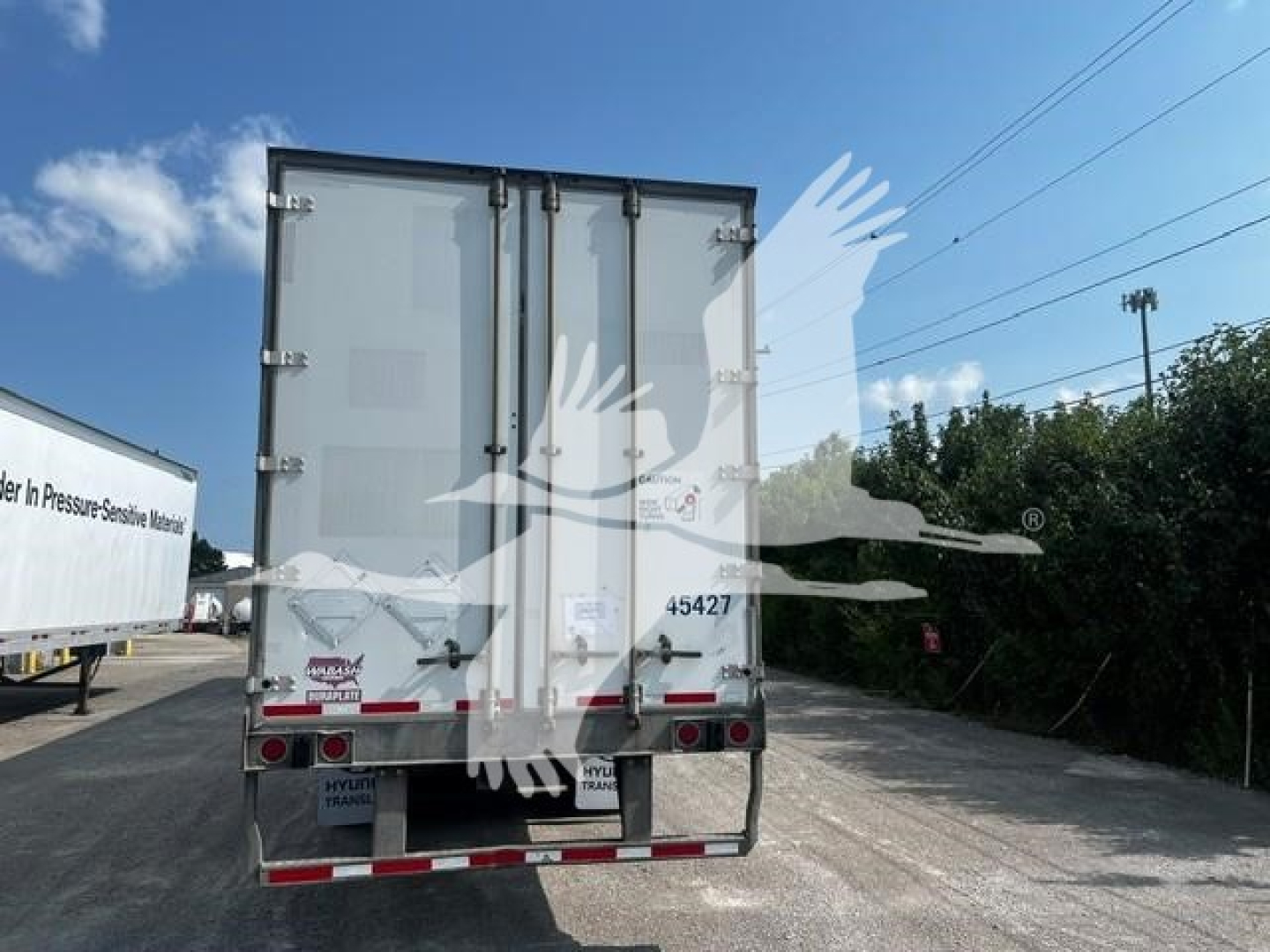 Rear view of a white enclosed trailer with double doors, red and white reflectors, and various stickers and license plate area.