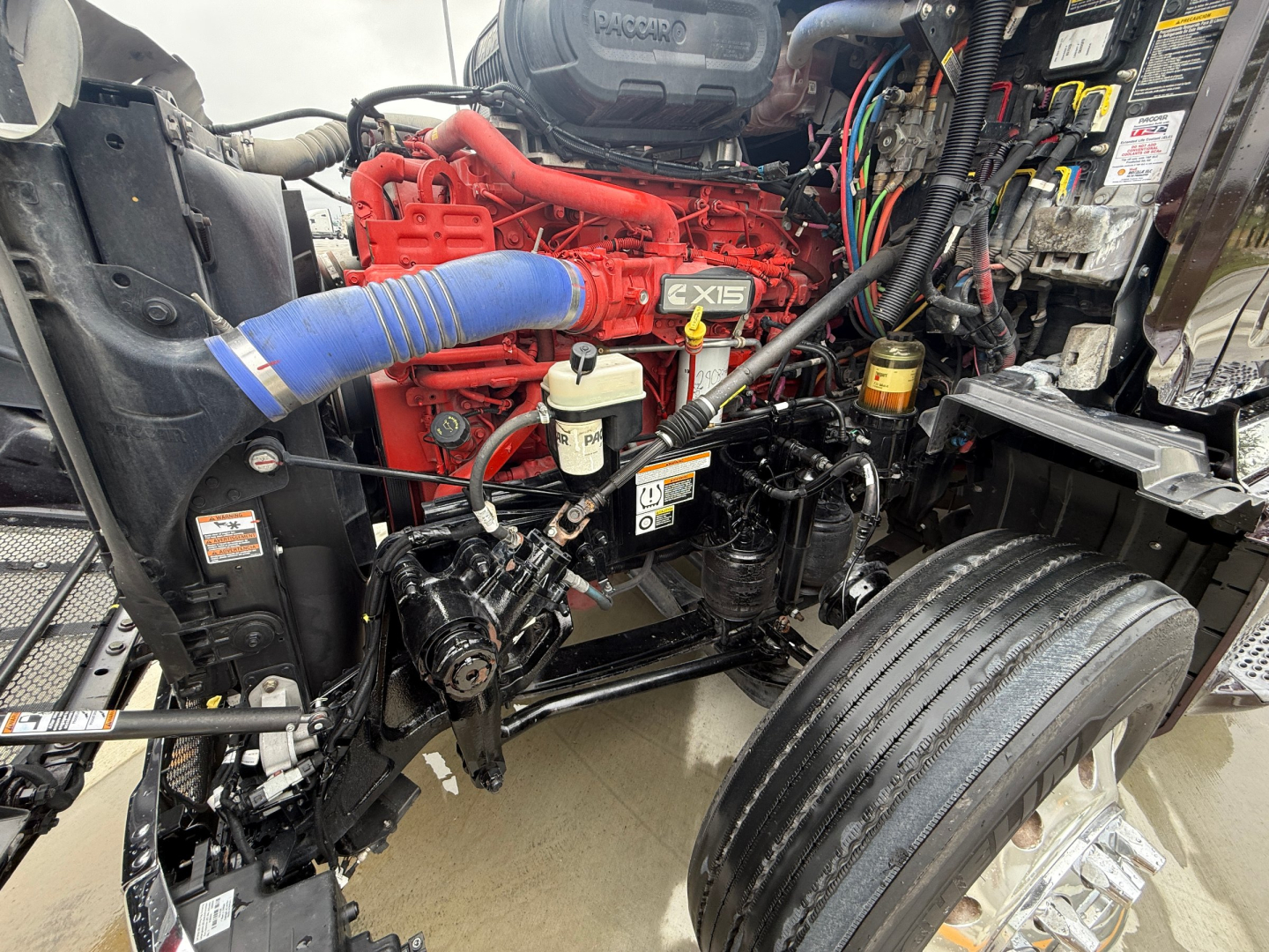 Peterbilt 579 engine bay with red engine and blue cold-air intake hose; visible belts, hoses, and wiring near front wheel well.