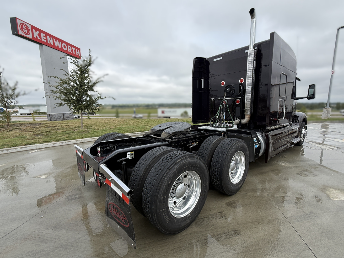 Peterbilt 579 tractor rear view showing dual rear axles and sleeper cab, black exterior, chrome exhaust stack, mud flaps, and tank area; Kenworth dealership in background.