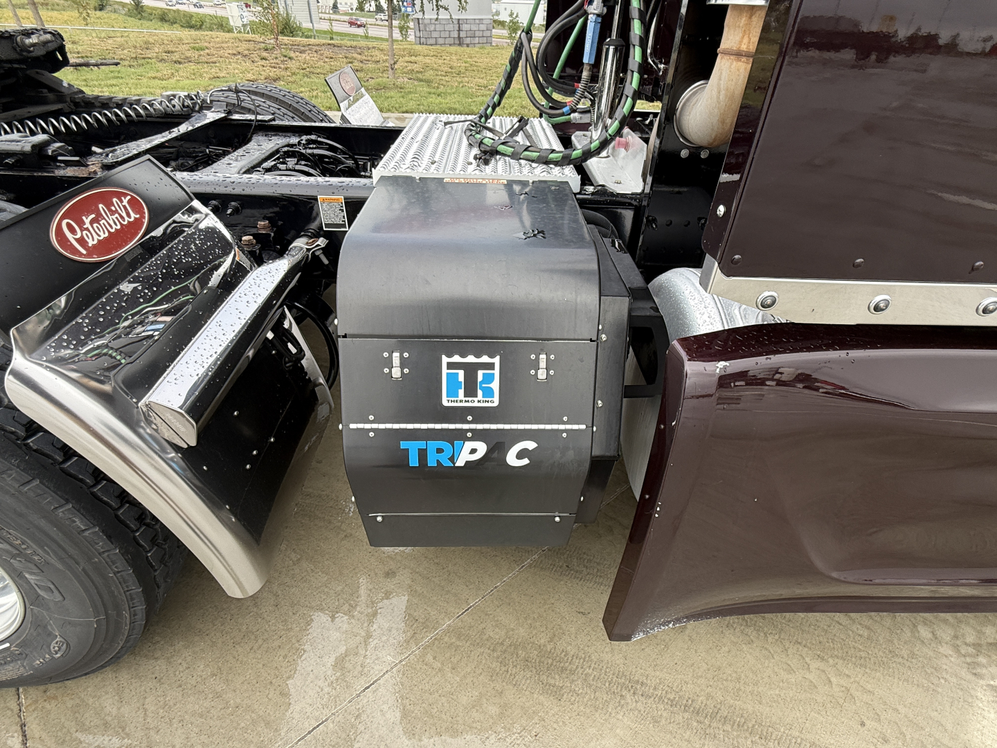 Truck cab area showing TRP C battery/box with Peterbilt logo emblem and nearby chrome fender; visible wheel and steps on left; maroon sleeper cab on right.