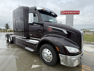 Peterbilt 579 conventional sleeper tractor in black, side view showing chrome bumper, large front grille, dual rear axles, and Kenworth dealership in background.
