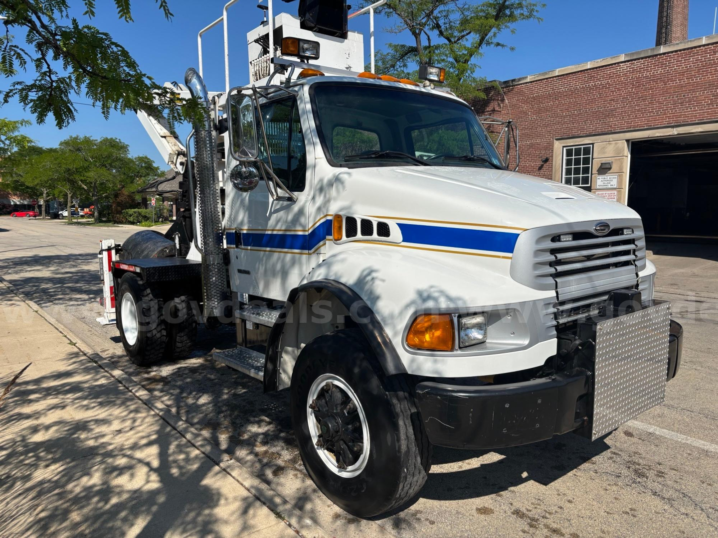 Grapple truck in-service showing front grille, bumper, and operator cab with blue and gold stripe; hydraulic boom controls on cab; windshield wipers; large side mirrors; white body.