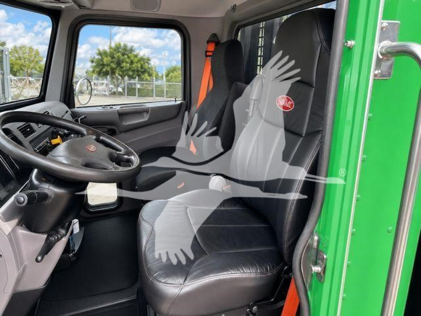Truck interior cab of a 2017 Peterbilt 348 showing black leather seat, steering wheel, and door with bright green exterior; seat has Peterbilt logo; window view of lot.