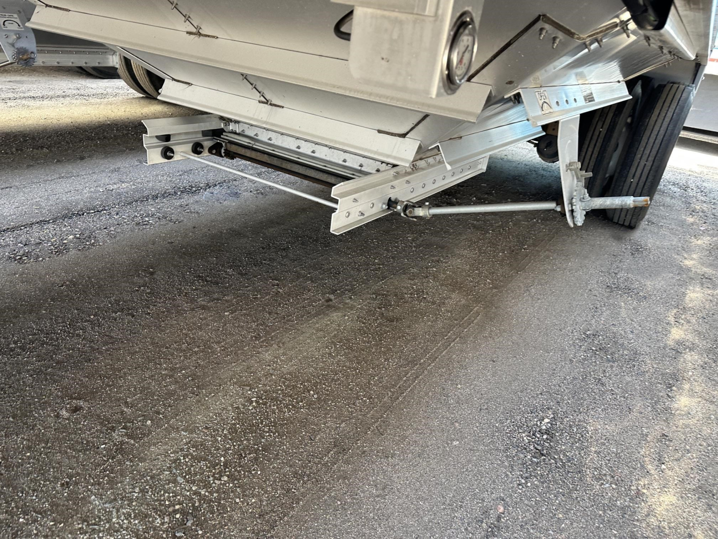 Trailer undercarriage showing metal frame, hydraulic/hatch support components, and wheel assembly. Visible metal rails and linkage near landing gear.