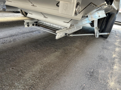 Trailer undercarriage showing metal frame, hydraulic/hatch support components, and wheel assembly. Visible metal rails and linkage near landing gear.