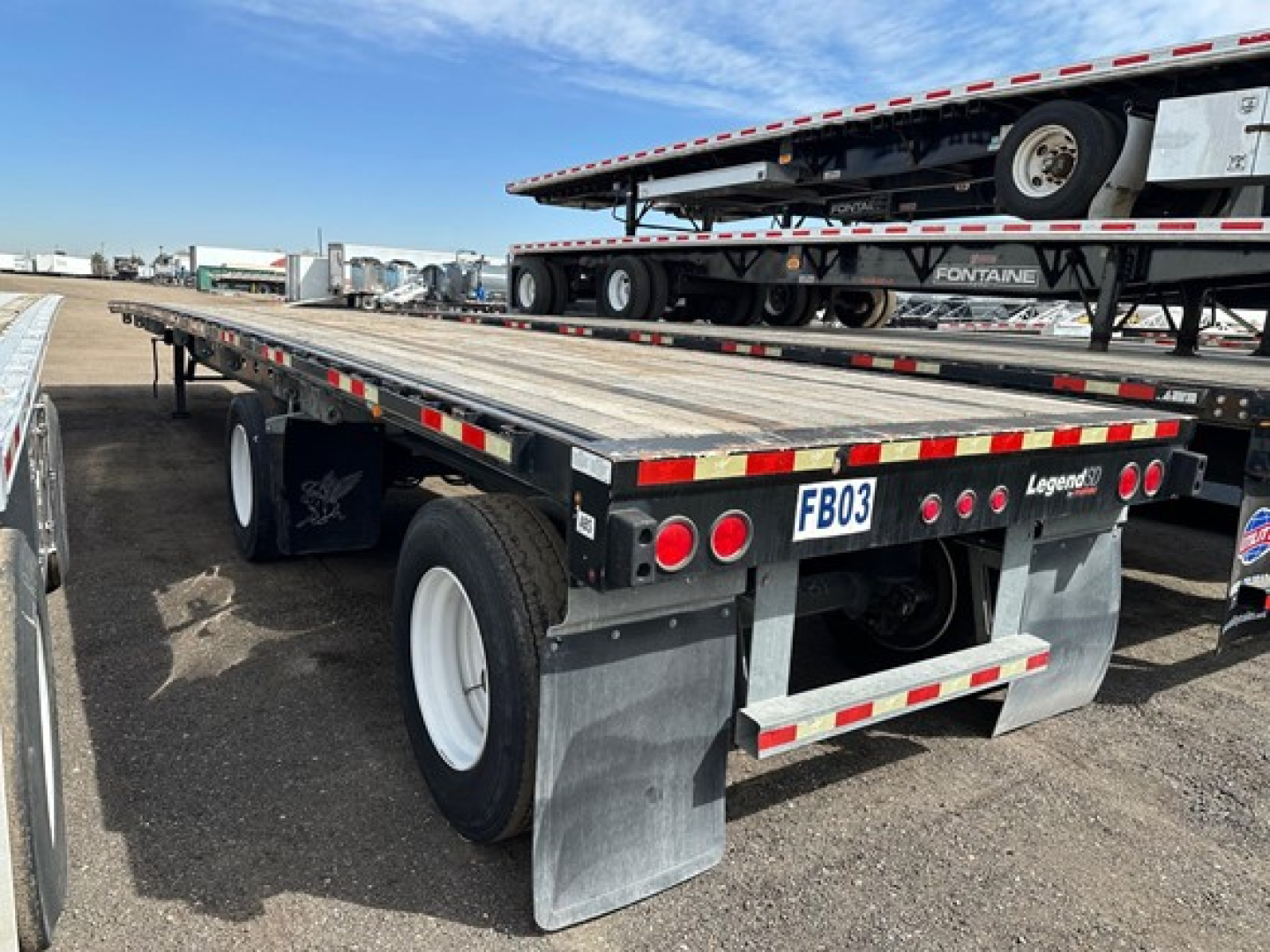 Flatbed trailer with dual rear axles and HD mudflaps, viewed from the left rear, showing reflective tape, license plate FB03, and a stacked other trailer in the background.