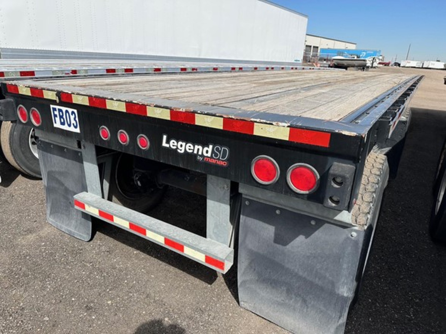 Manac flatbed trailer rear view showing deck, mud flaps, and red/white reflective tape with FB03 license plate area and Legend SD branding.