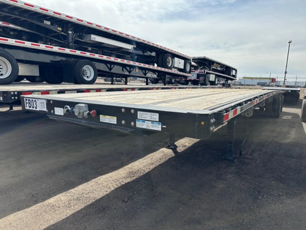 Flatbed trailer showing the deck and undercarriage with ramps stowed, edge rails, and wheelset. Visible labels and signage on the front edge and side.