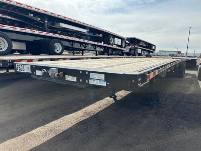 Flatbed trailer showing the deck and undercarriage with ramps stowed, edge rails, and wheelset. Visible labels and signage on the front edge and side.