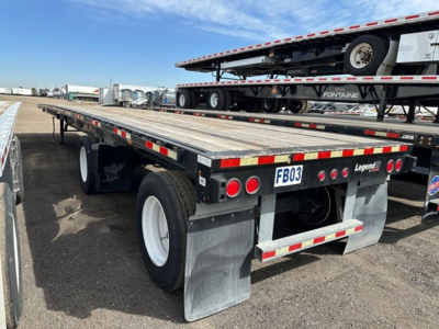 Flatbed trailer with dual rear axles and HD mudflaps, viewed from the left rear, showing reflective tape, license plate FB03, and a stacked other trailer in the background.