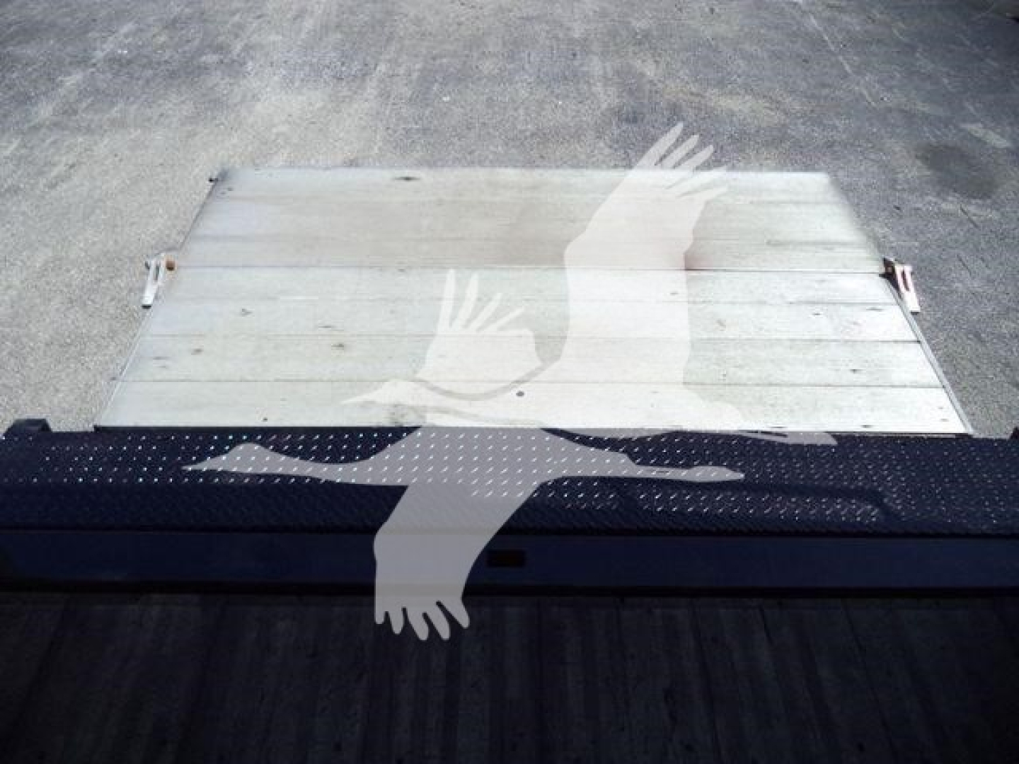 Flatbed truck bed with a metallic silver aluminum surface and a black perforated step looking at the rear of a Hino 195 cab; no legible text visible.