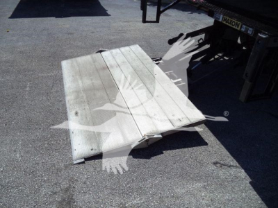 Flat aluminum loading ramp or platform with ribbed surface lying on pavement, attached to a truck trailer at rear.