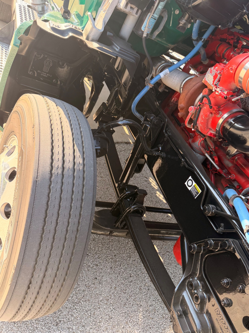 Kenworth T880 front wheel and engine bay components showing front axle, suspension, red engine block, hoses, and blue wiring.