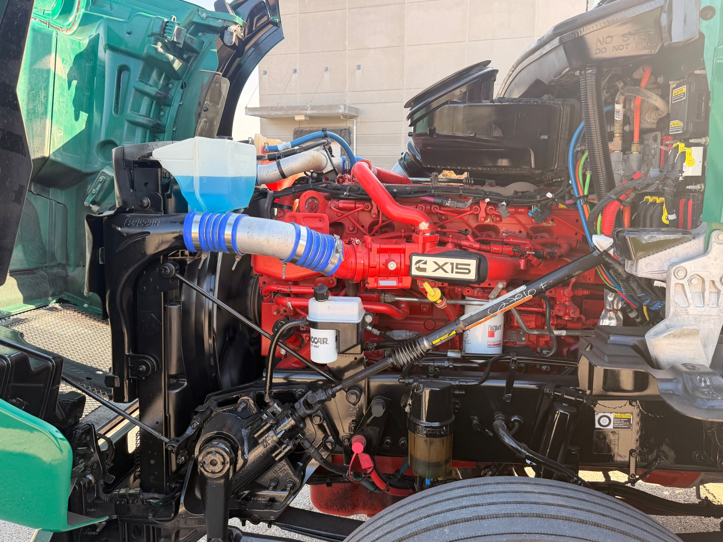 Kenworth T880 engine bay with red X15 engine and visible blue hoses, coolant reservoir, and various hoses and wiring in engine compartment.