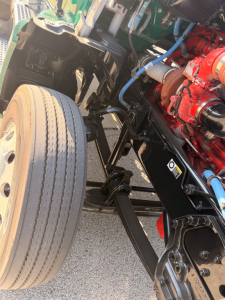 Kenworth T880 front wheel and engine bay components showing front axle, suspension, red engine block, hoses, and blue wiring.