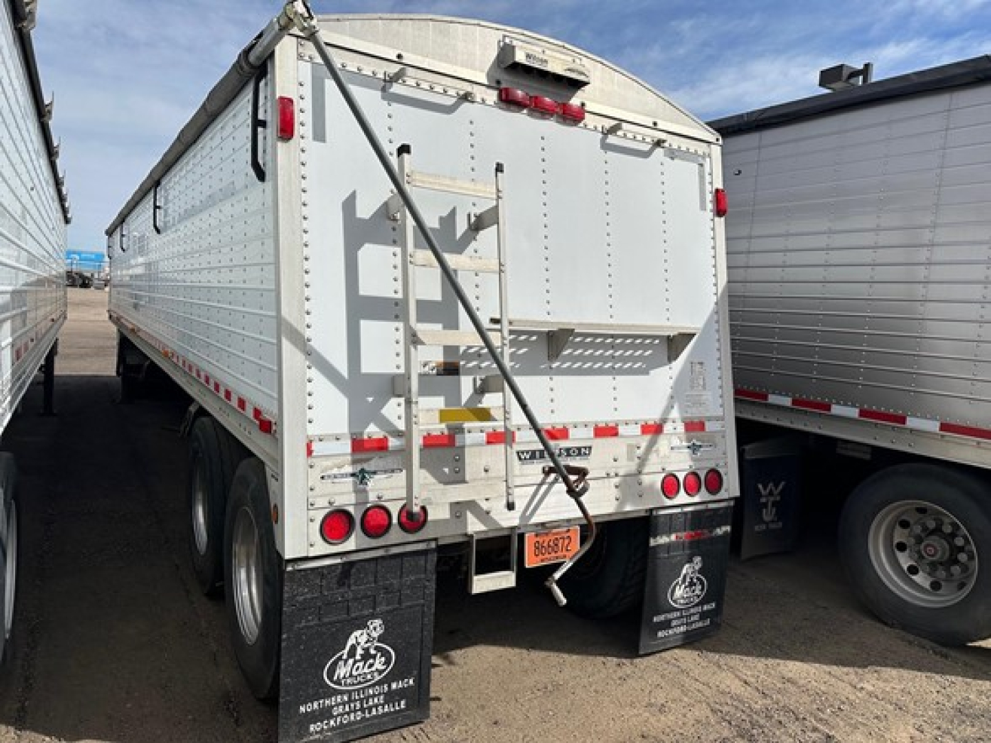 Trailer rear showing hopper grain trailer with tailgate, vertical support brace, rear lights, Mud flaps, and license plate; white aluminum panels; WVU sticker and branding.