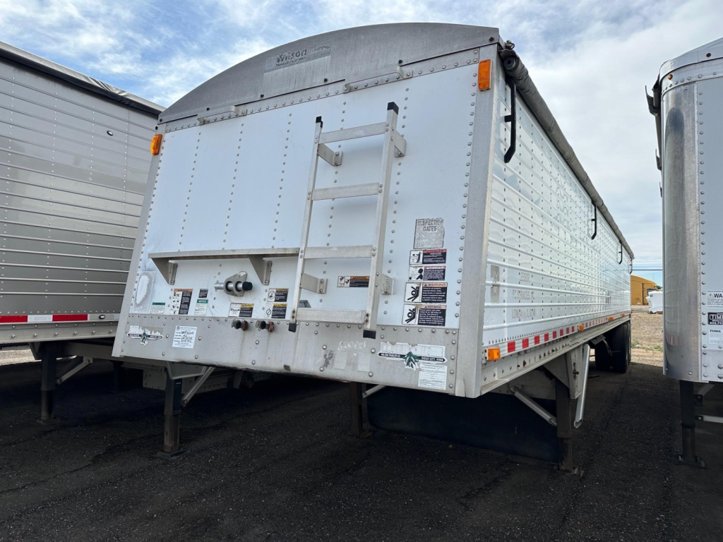 Trailer rear showing 2008 Wilson grain hopper trailer with ladder, rear doors, and warning labels. VIN sticker and stickers on left door surrounded by riveted aluminum panels.