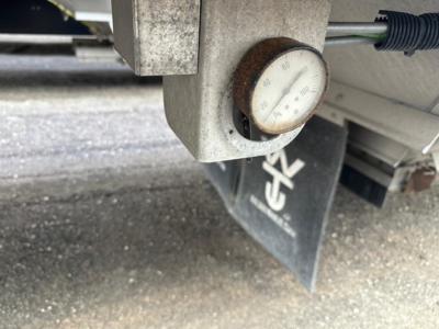 Gauge and metal bracket on a Wilson grain trailer showing a round dial gauge with numbers and a rubber-bushed pipe nearby.