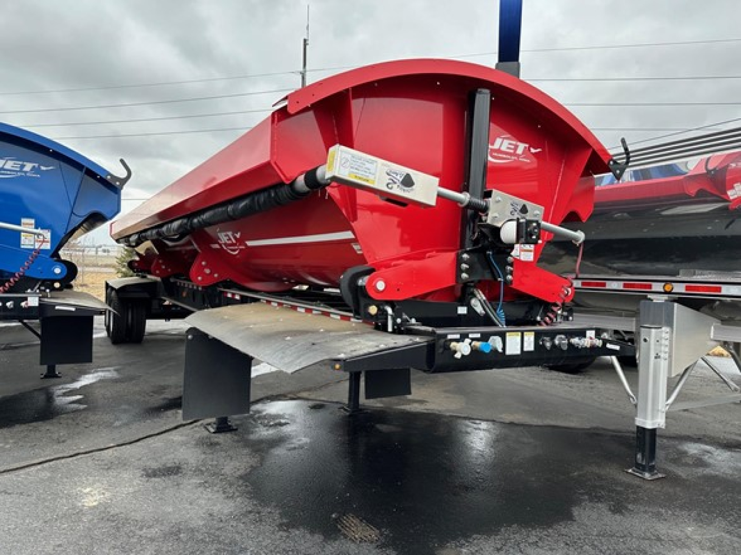 Jet Jet side dump trailer showing red steel hopper and hydraulic hoist with control valve and hitch; trailer on a stand with black base and gray platform.