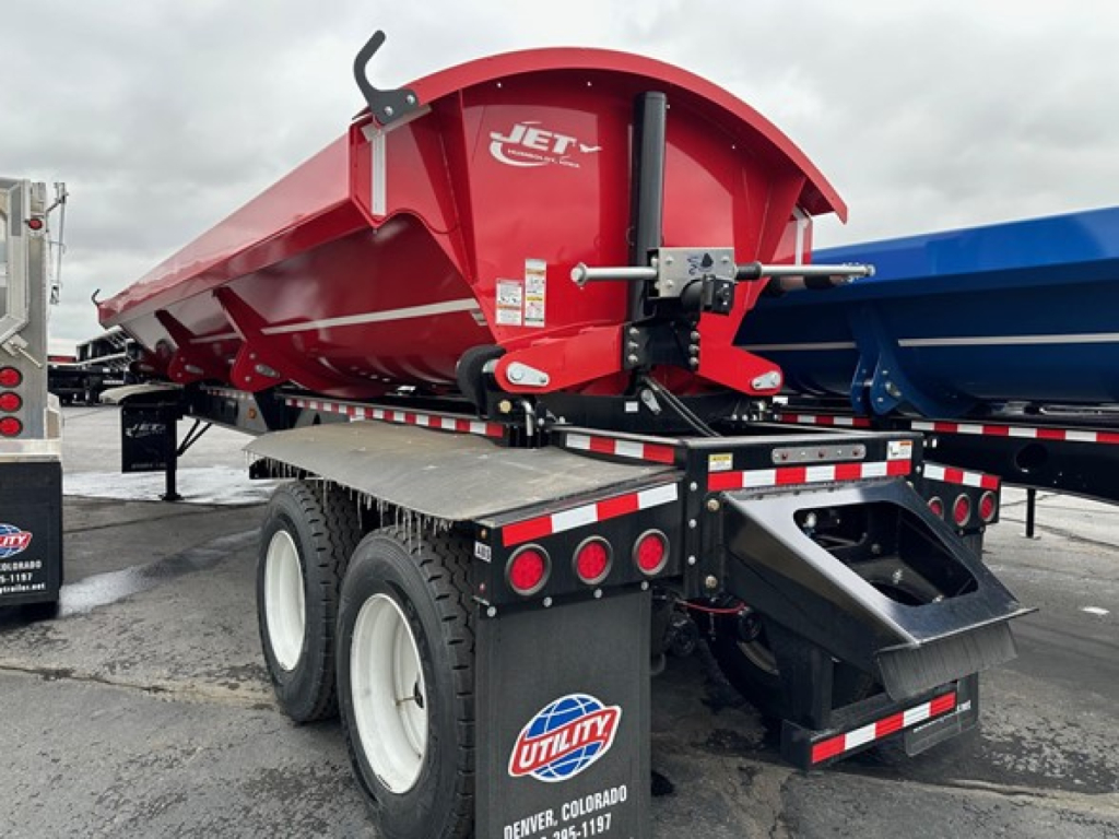 Red Jet side dump trailer viewed from rear-left, showing dump bed with Jet logo, hydraulic cylinder, wheels, and reflective safety bars.