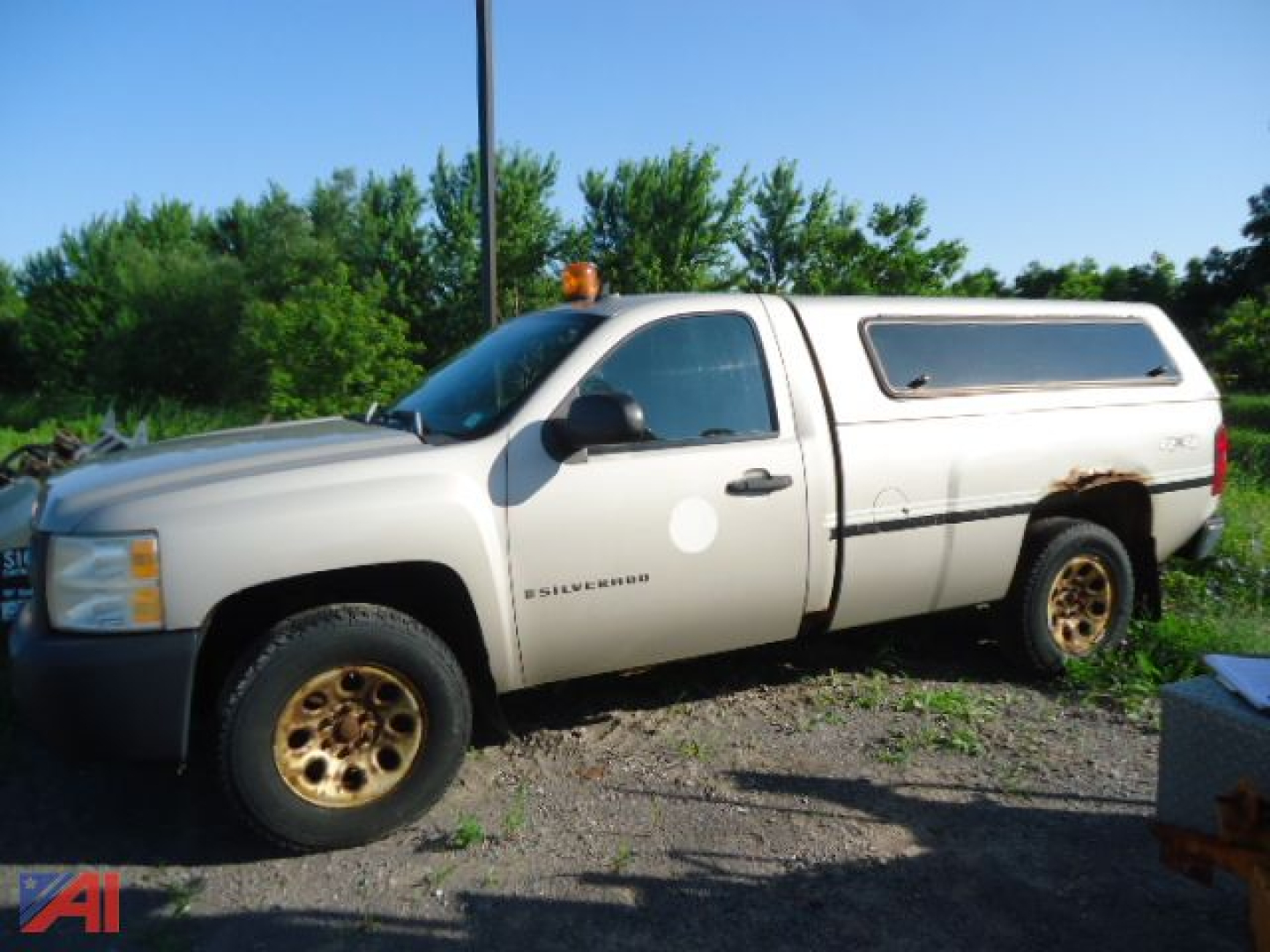 Chevrolet Silverado 1500 pickup with camper shell and orange beacon light on roof, front-left view, beige color, wheel rims, in-service condition.
