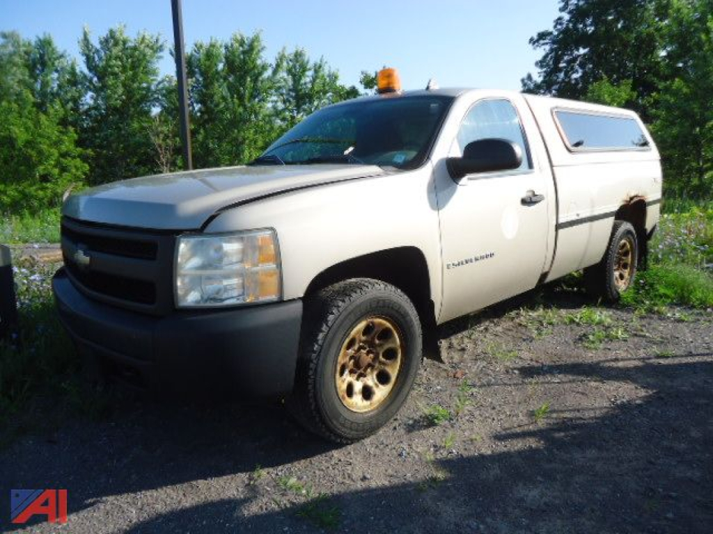 Chevrolet Silverado 1500 pickup truck in-service front-left view with utility camper cap, amber light on cab, beige body, black bumper and gold steel wheels.