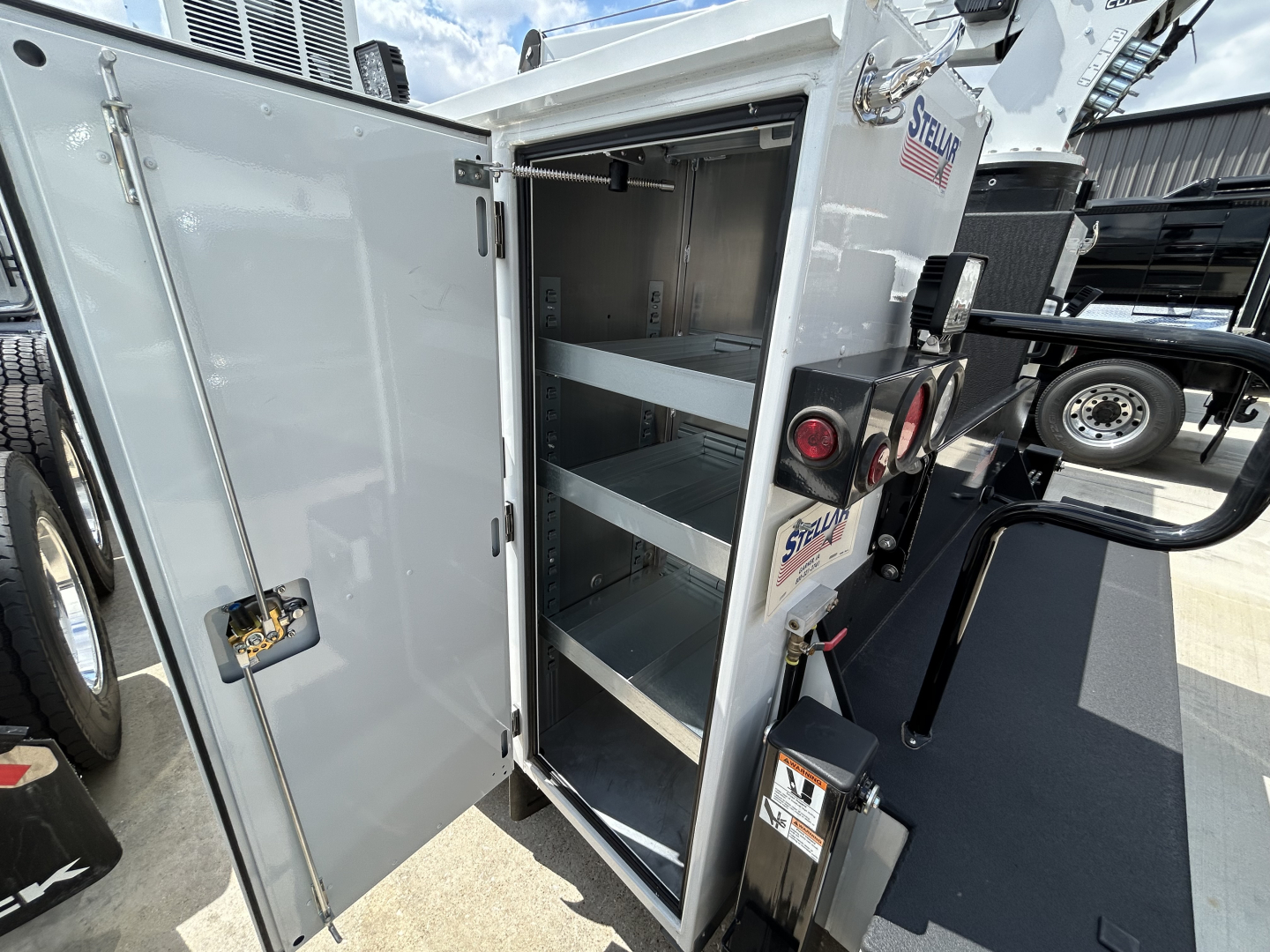 Service body vanes open showing shelving in compartment of 2025 Mack MD6 truck, with Spare/utility storage cabinet doors and hydraulic lift components visible.