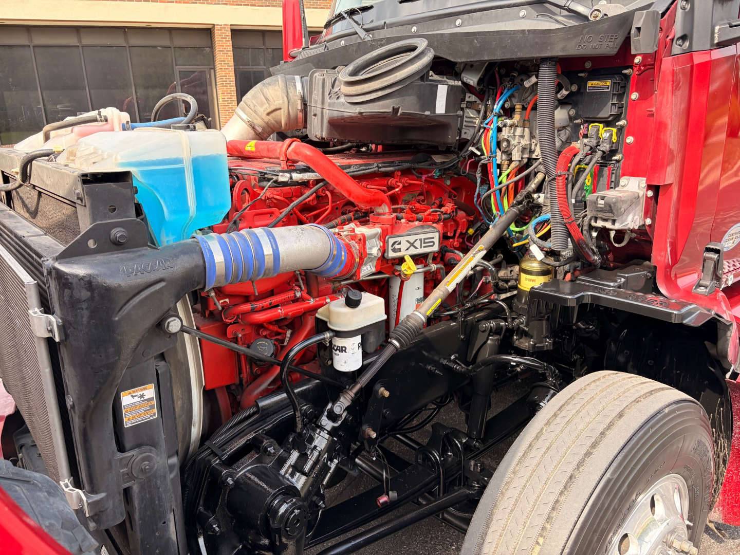 Engine bay of a 2022 Kenworth T680 showing red engine with EXIS badge, blue coolant reservoir, hoses, wiring, and front wheel well.