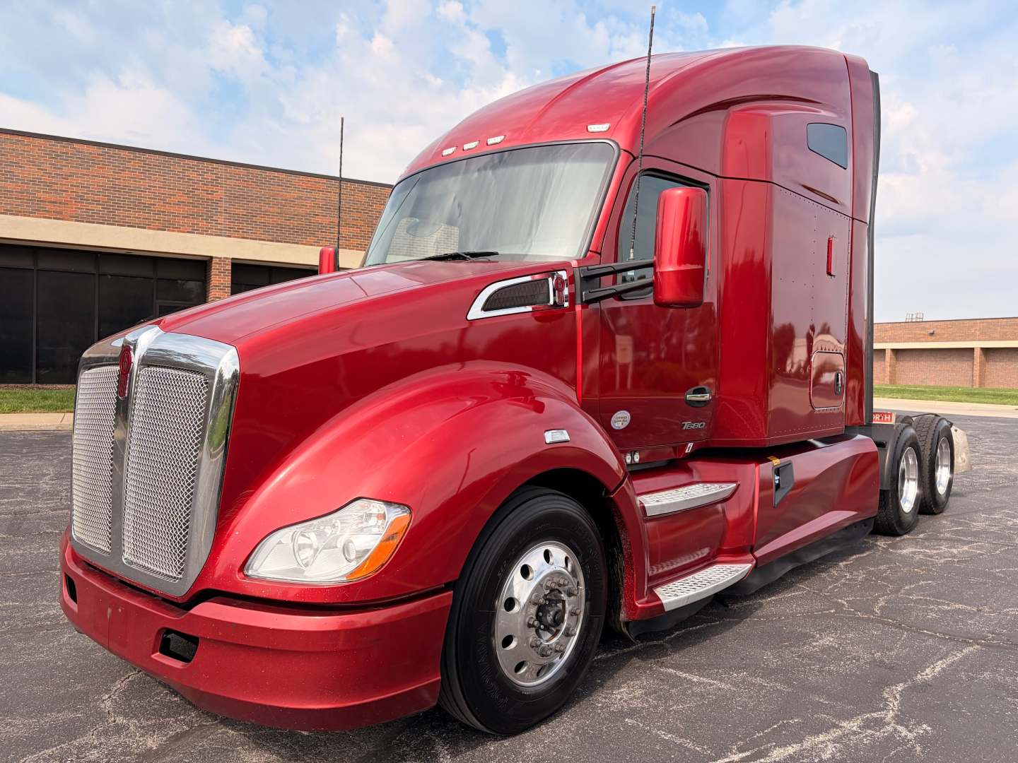 Red 2022 Kenworth T680 conventional sleeper truck in a parking lot, side view showing front grille, cab, and dual rear axles.