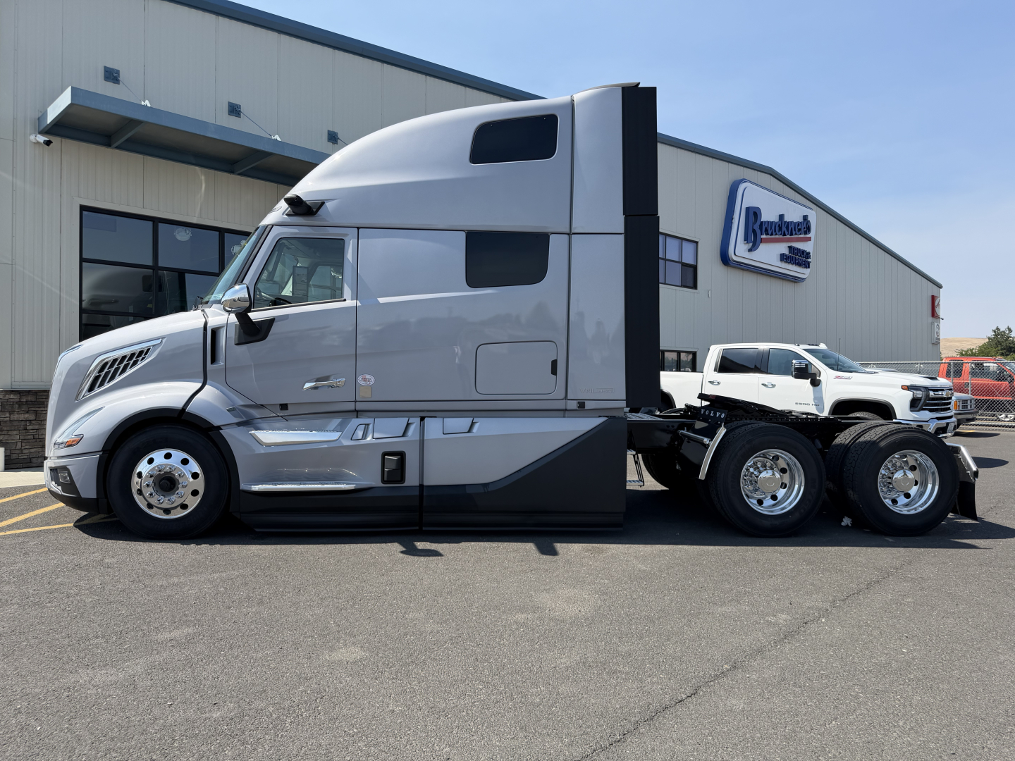 Truck cabin side view with a sleek gray Volvo VNL sleeper cab and chrome wheels, parked in front of a dealership building.