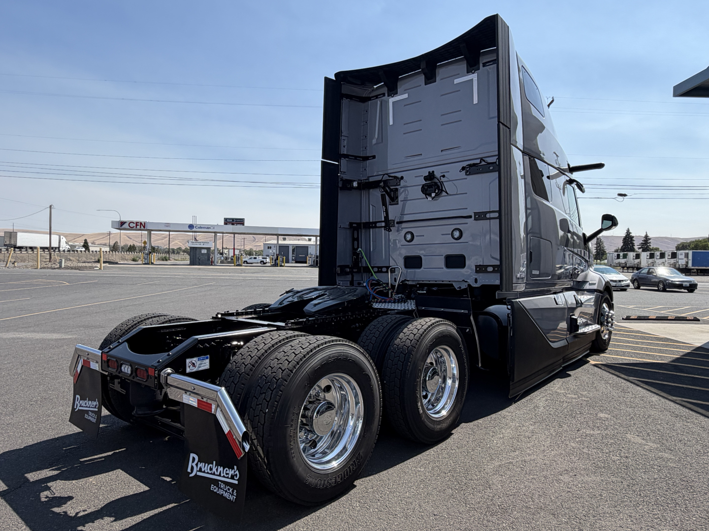 Side view of a new 2027 Volvo VNL64T860 conventional sleeper truck in a parking lot, showing triple rear axles, chrome wheels, and rear cab area.