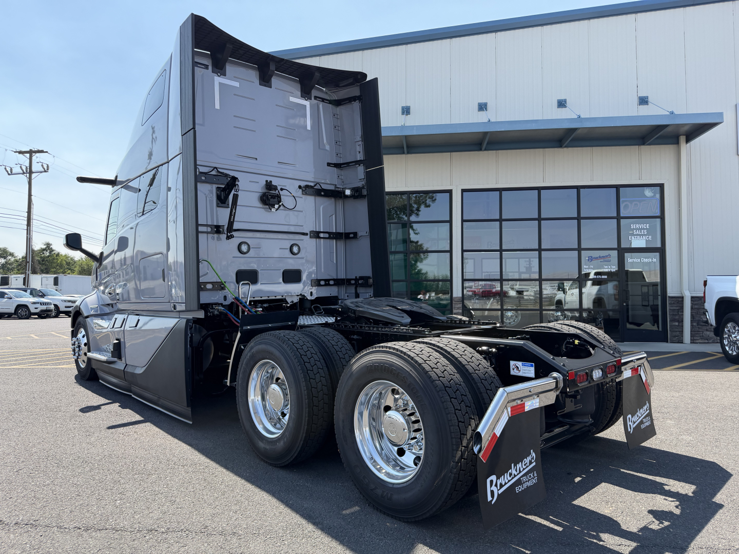 Truck rear view showing Volvo VNL sleeper cab with tandem rear axles and mud flaps; no visible text on vehicle. The background shows a dealership building with glass doors.