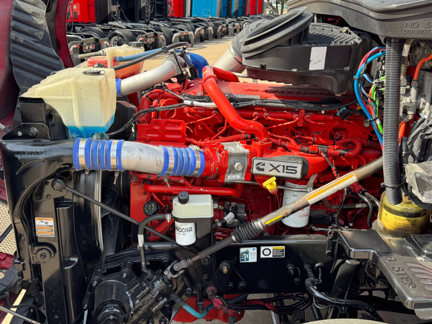 Kenworth T680 engine bay with red 12.0L engine, coolant reservoir, hoses, and various components visible