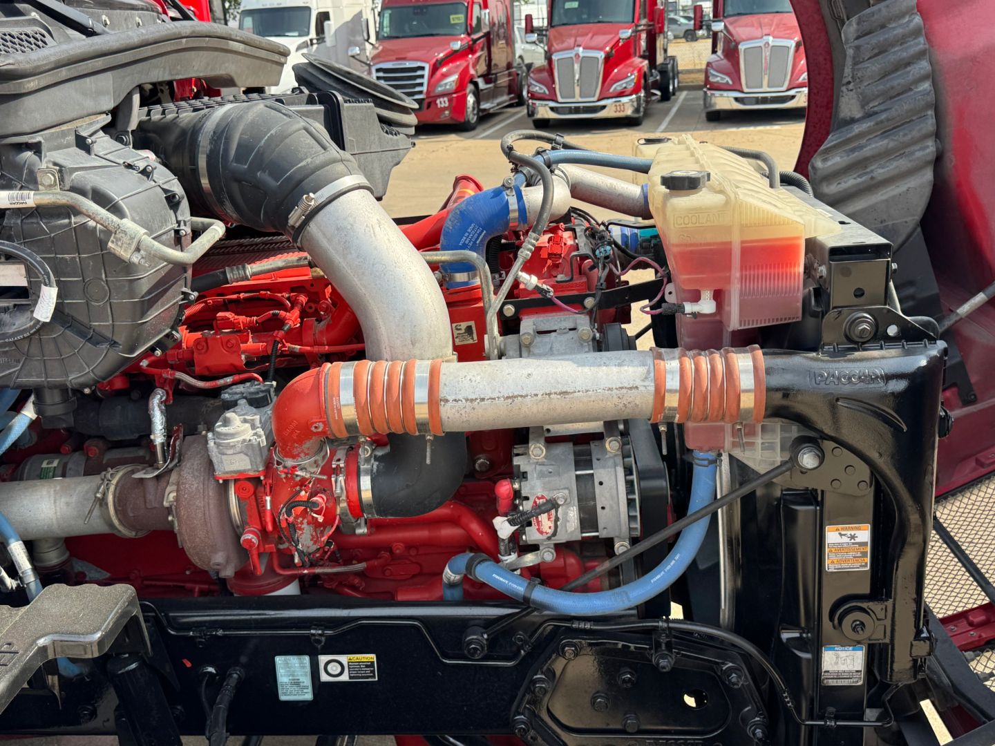 Kenworth T680 engine bay showing red engine, intercooler piping, coolant reservoir, and battery on right side.