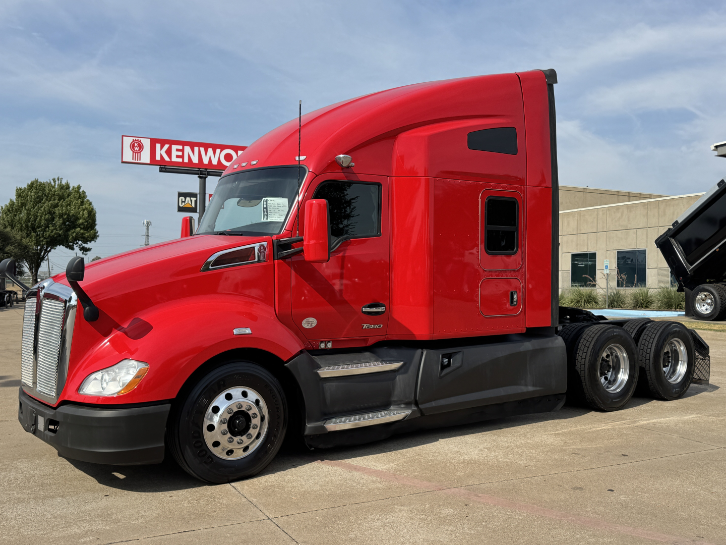 Kenworth T680 sleeper cab tractor in red, side view, with Kenworth dealership in background.