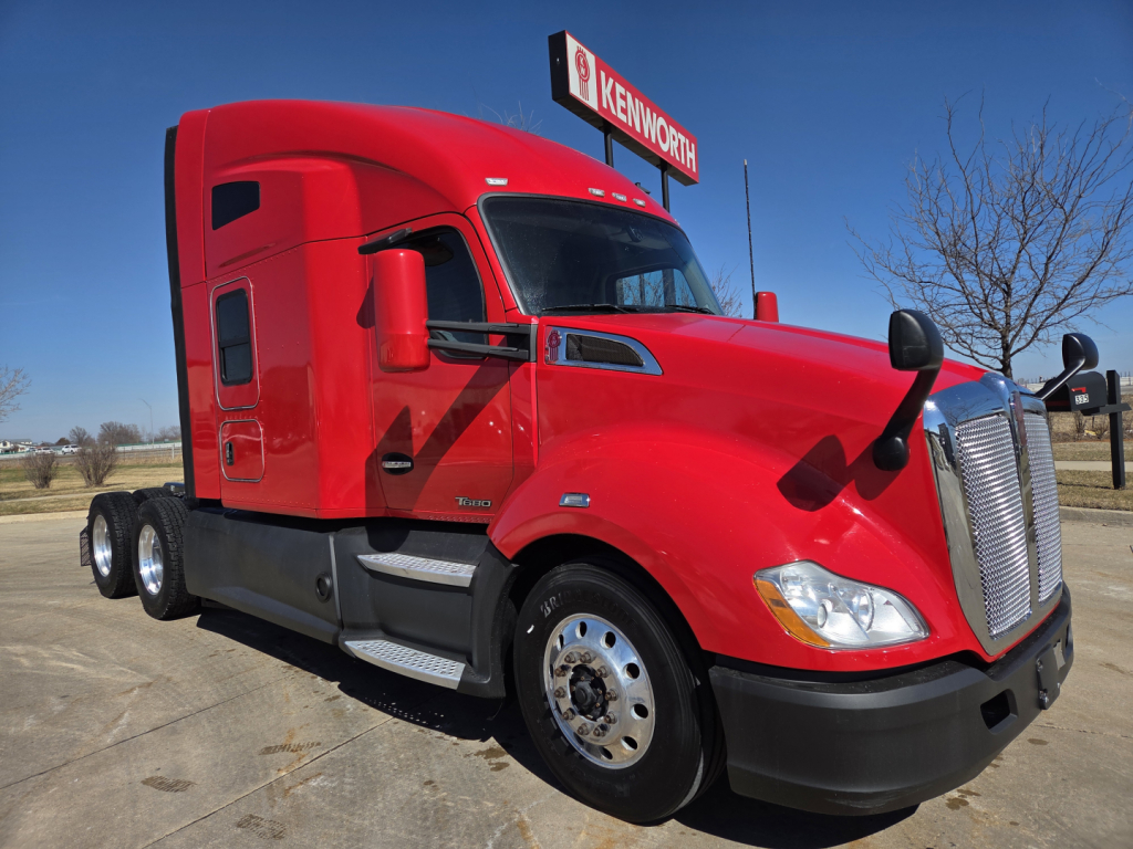 Kenworth T680 cab-over semi-truck in red, front three-quarter view, shows sleeper cab, dual rear axles, chrome grille, and Kenworth sign in background.