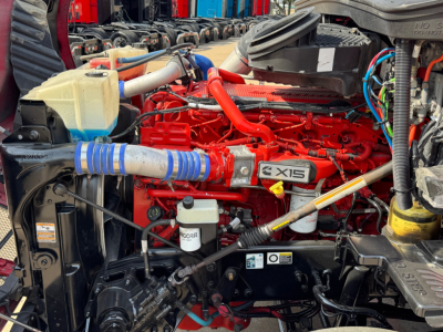 Kenworth T680 engine bay with red 12.0L engine, coolant reservoir, hoses, and various components visible