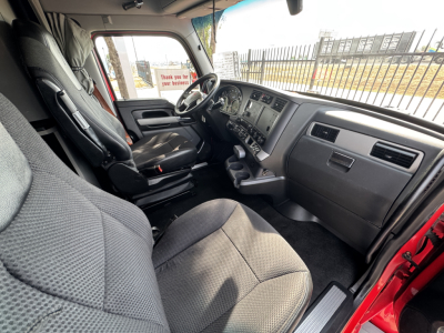 Kenworth T680 interior cab view with driver seat and dashboard, steering wheel, center console, and passenger side; gray cloth seats; door open showing outside fence.