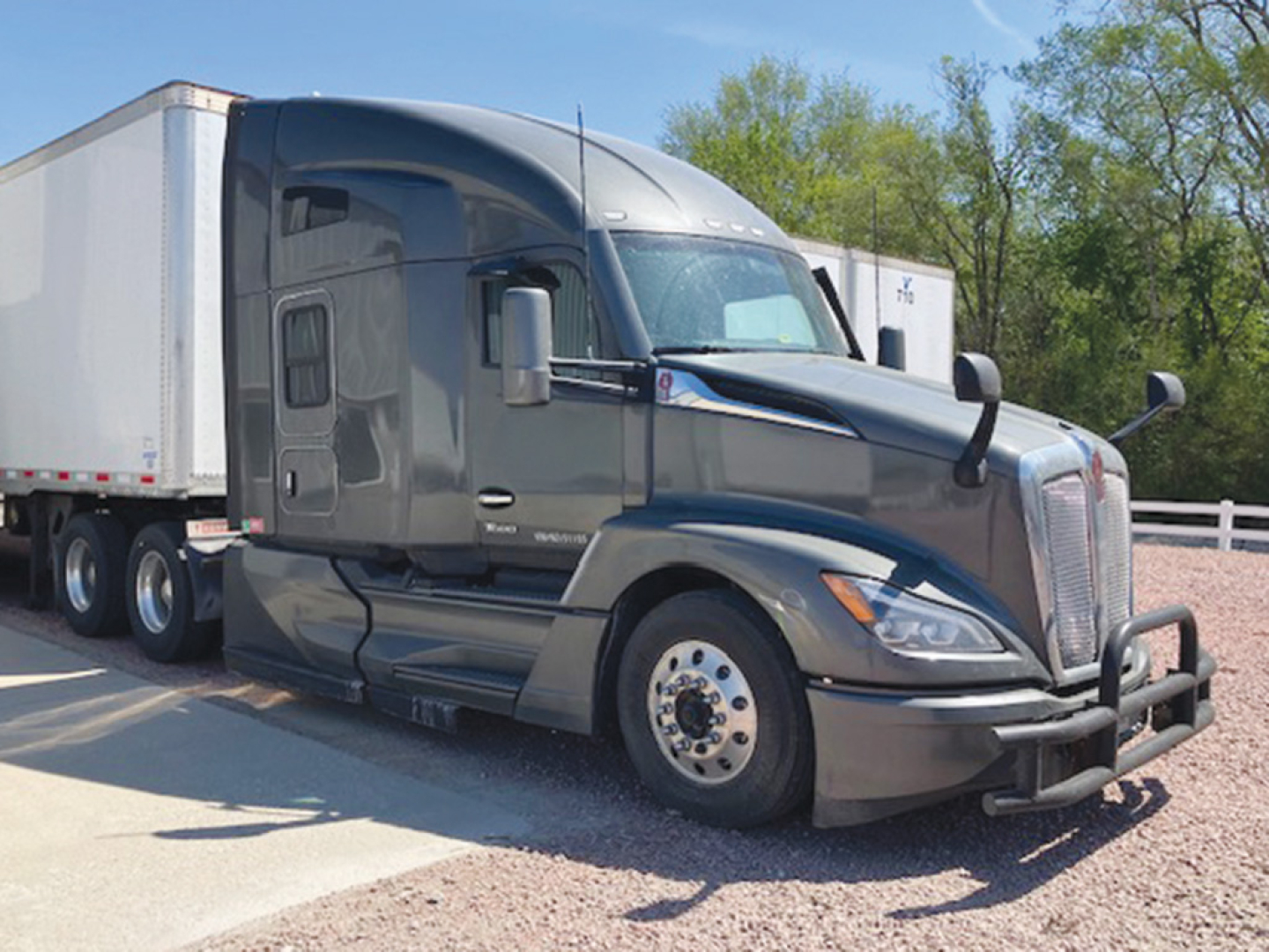 Kenworth T680 Next Gen tractor with a large chrome grille, gray paint, side mirrors, aero cab, and attached white trailer visible.