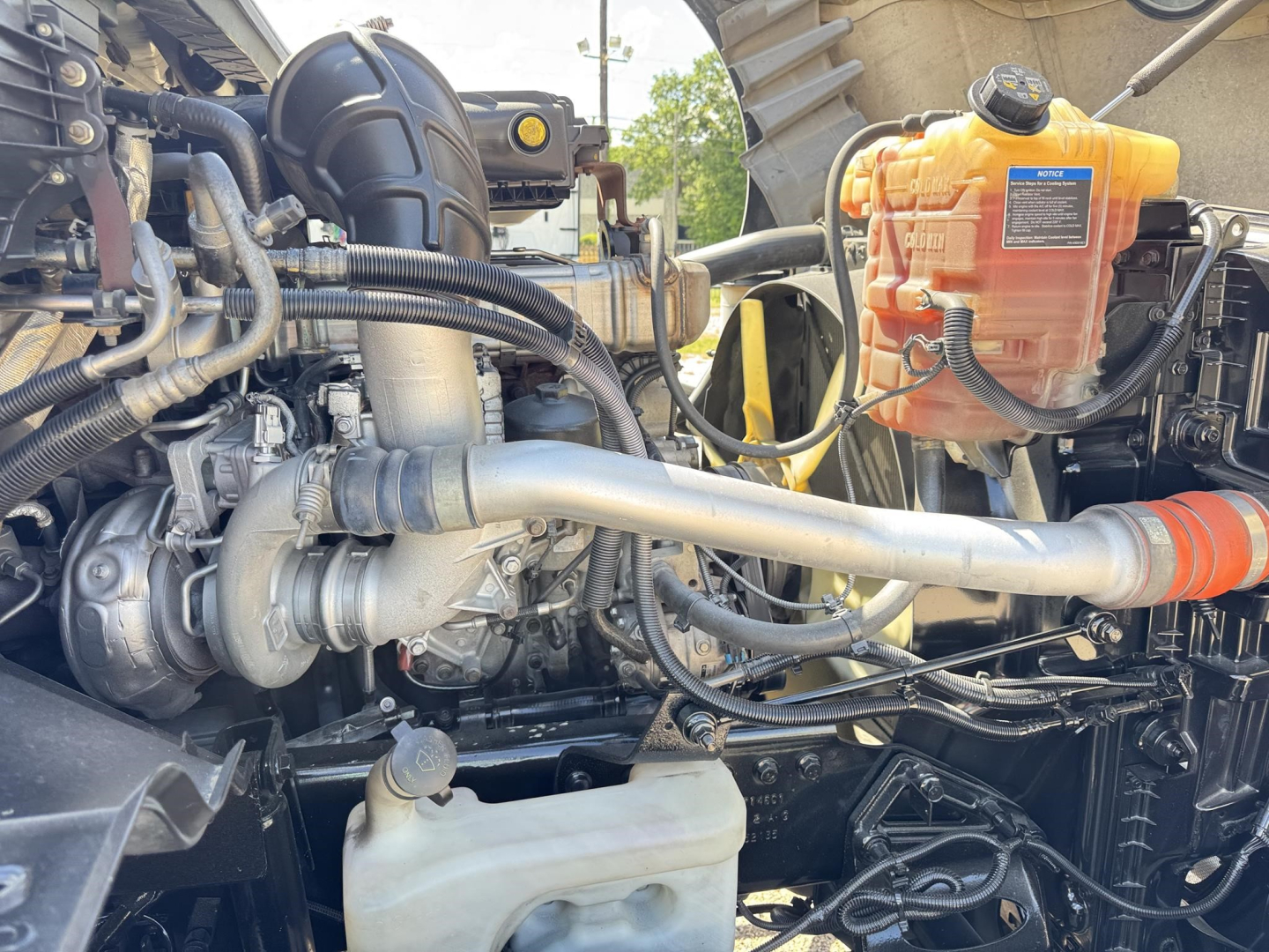 Truck engine bay view with turbocharger, hoses, and coolant reservoir labeled; orange coolant reservoir on the right with blue cap; various metallic pipes and wiring visible.