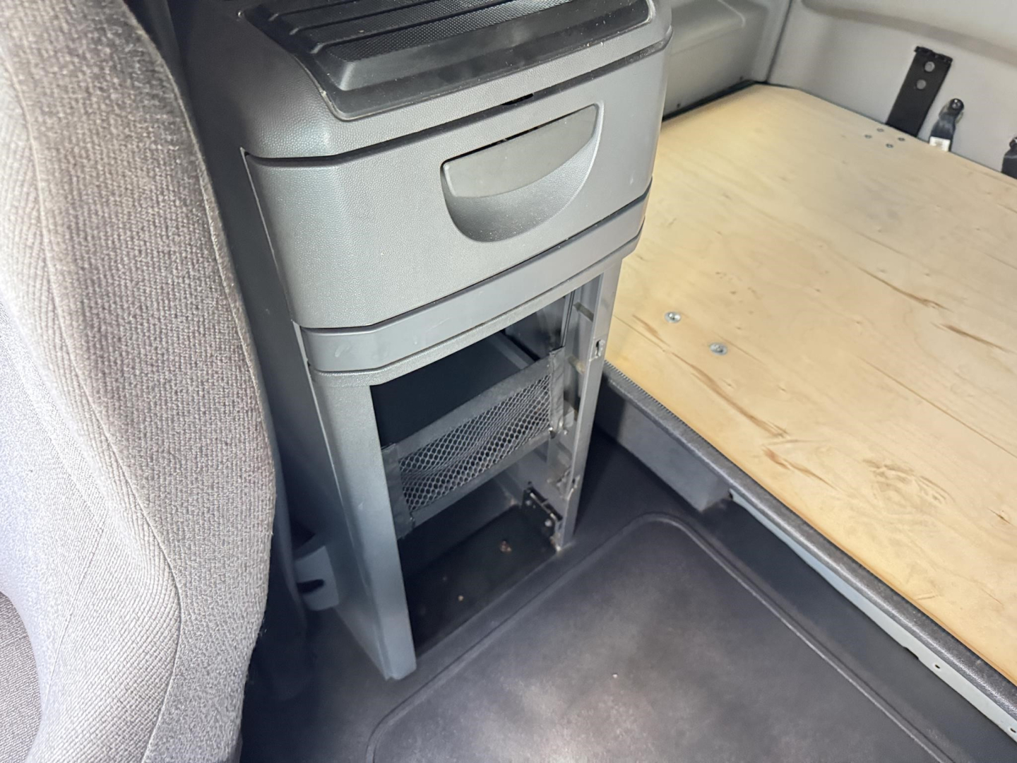 Truck interior storage area under sleeper with open cabinet and sliding drawer, wood board surface, and metal panels visible.