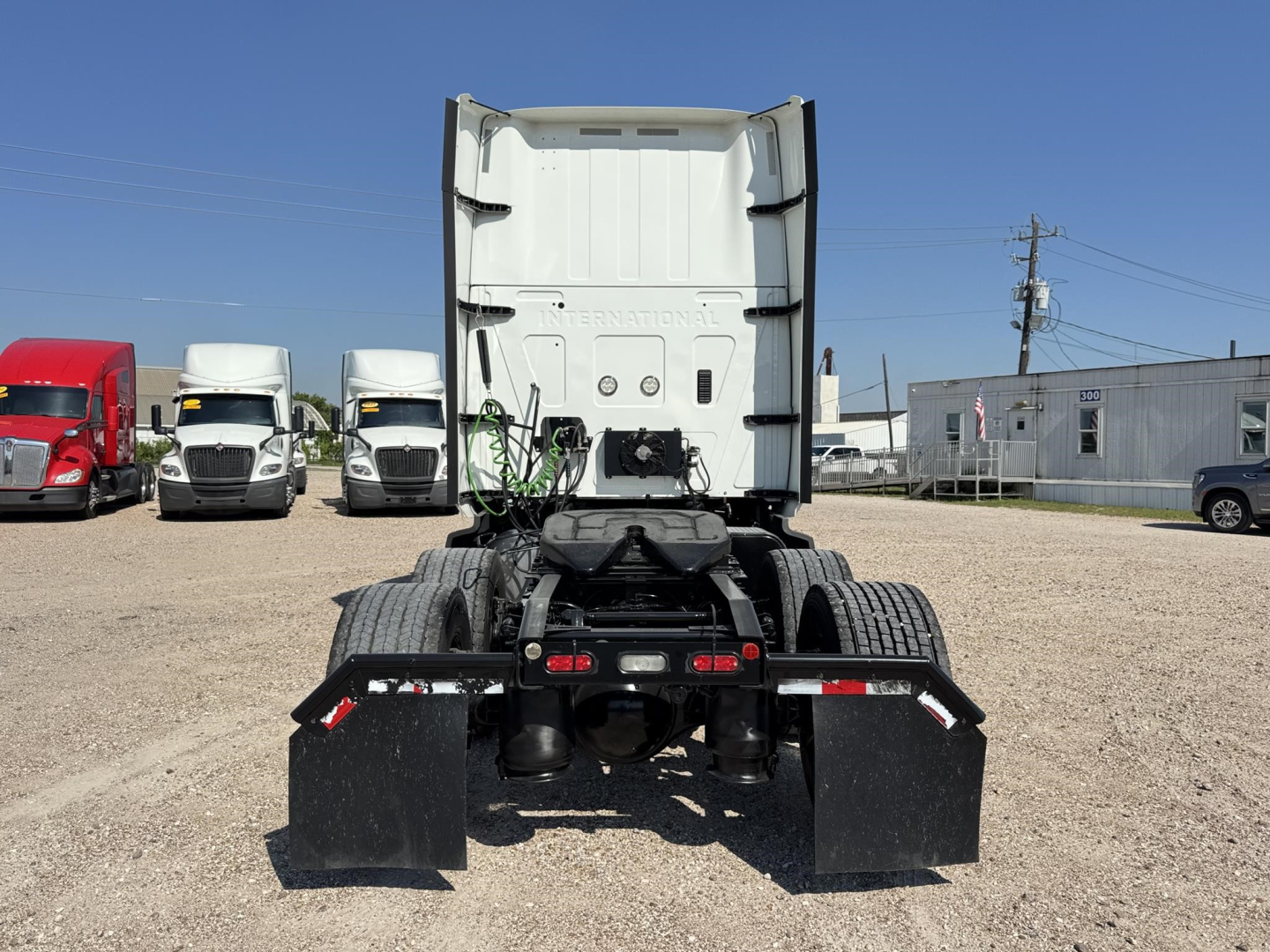 Truck cab and chassis rear view showing fifth wheel, mud flaps, and dual rear tires; white sleeper cab back with back-of-cab components visible; several trucks parked in background.