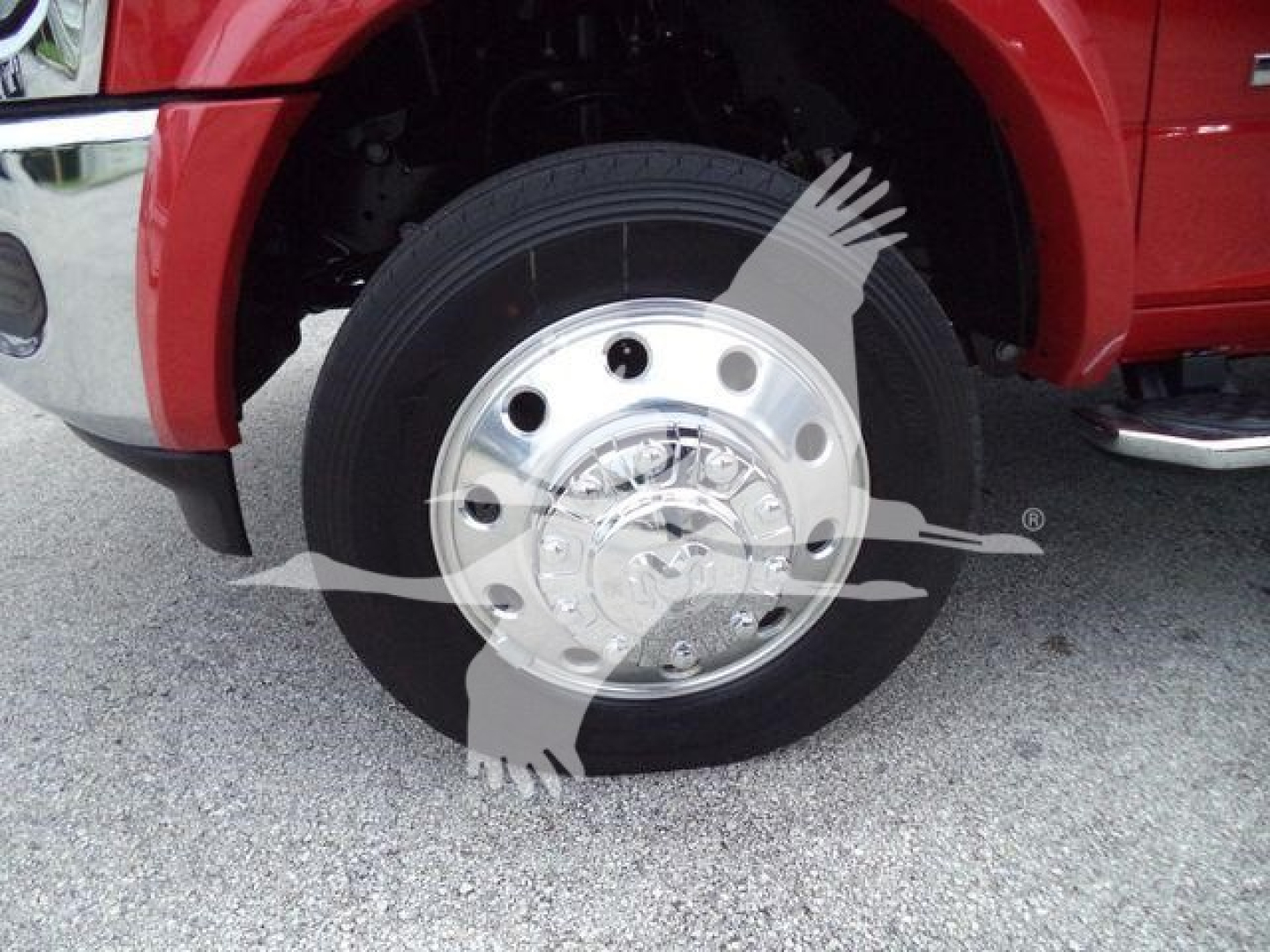 Front wheel and hubcap of a red Ram truck showing large chrome wheel with black tire and partial fender; watermark with a gloved hand and wrench overlay.