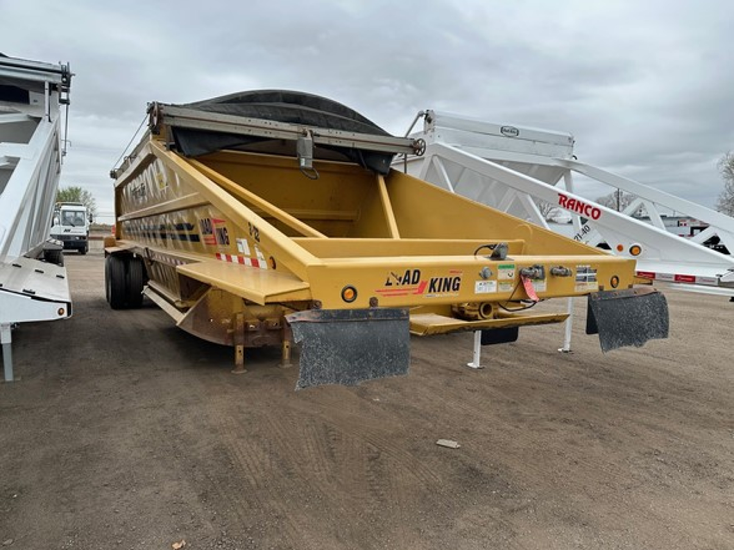 Load King Originator bottom dump trailer in yellow with rear tailgate and ramps, side rails, and spare wheel on top; visible chain hooks and warning decals.
