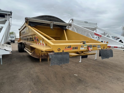 Load King Originator bottom dump trailer in yellow with rear tailgate and ramps, side rails, and spare wheel on top; visible chain hooks and warning decals.