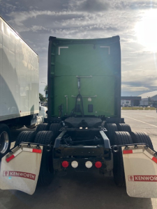 Kenworth T680 rear view showing green sleeper cab, dual rear axles, mud flaps with Kenworth logos, and trailer hitch hardware.