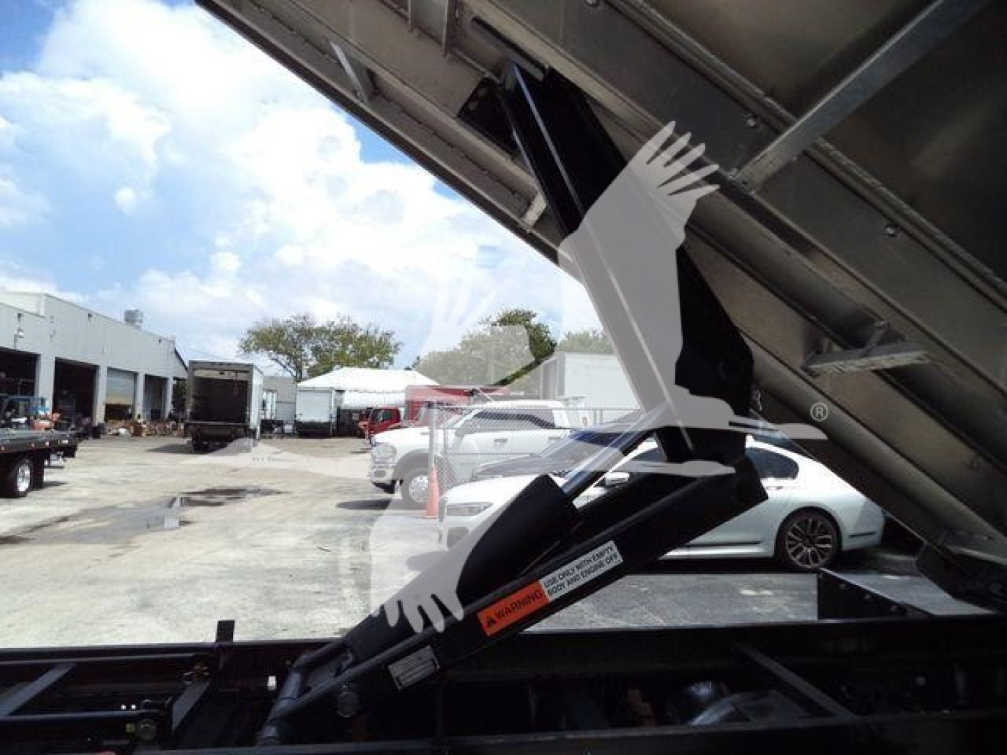 Ford F-550 dump bed side view showing hydraulic cylinder and pulley mechanism in raised position with surrounding lot and vehicles.
