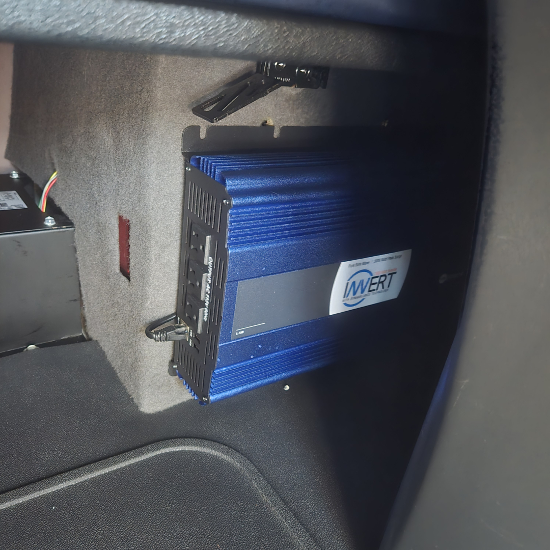 In-vehicle view showing a blue inverter/Power Control Module mounted inside a sleeper cab area with wiring harness and bracket.