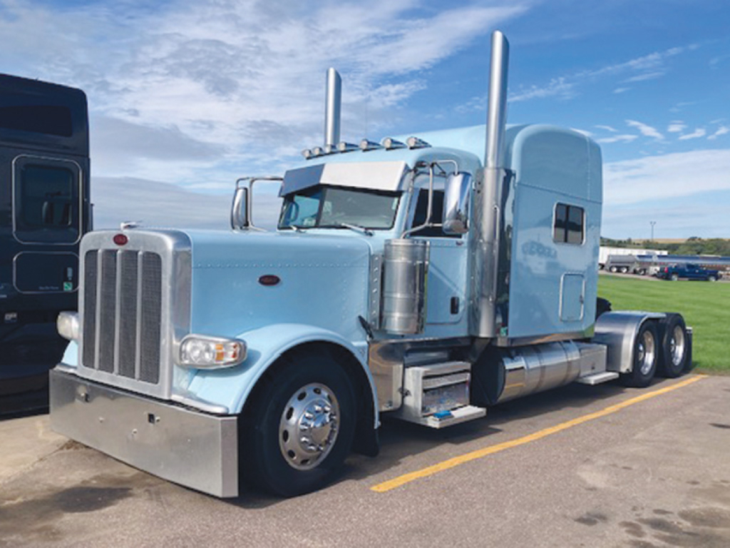 Peterbilt 389 UltraCab Extended Hood truck in blue, front-left view showing grille, dual exhaust stacks, chrome bumper, and sleeper cab.